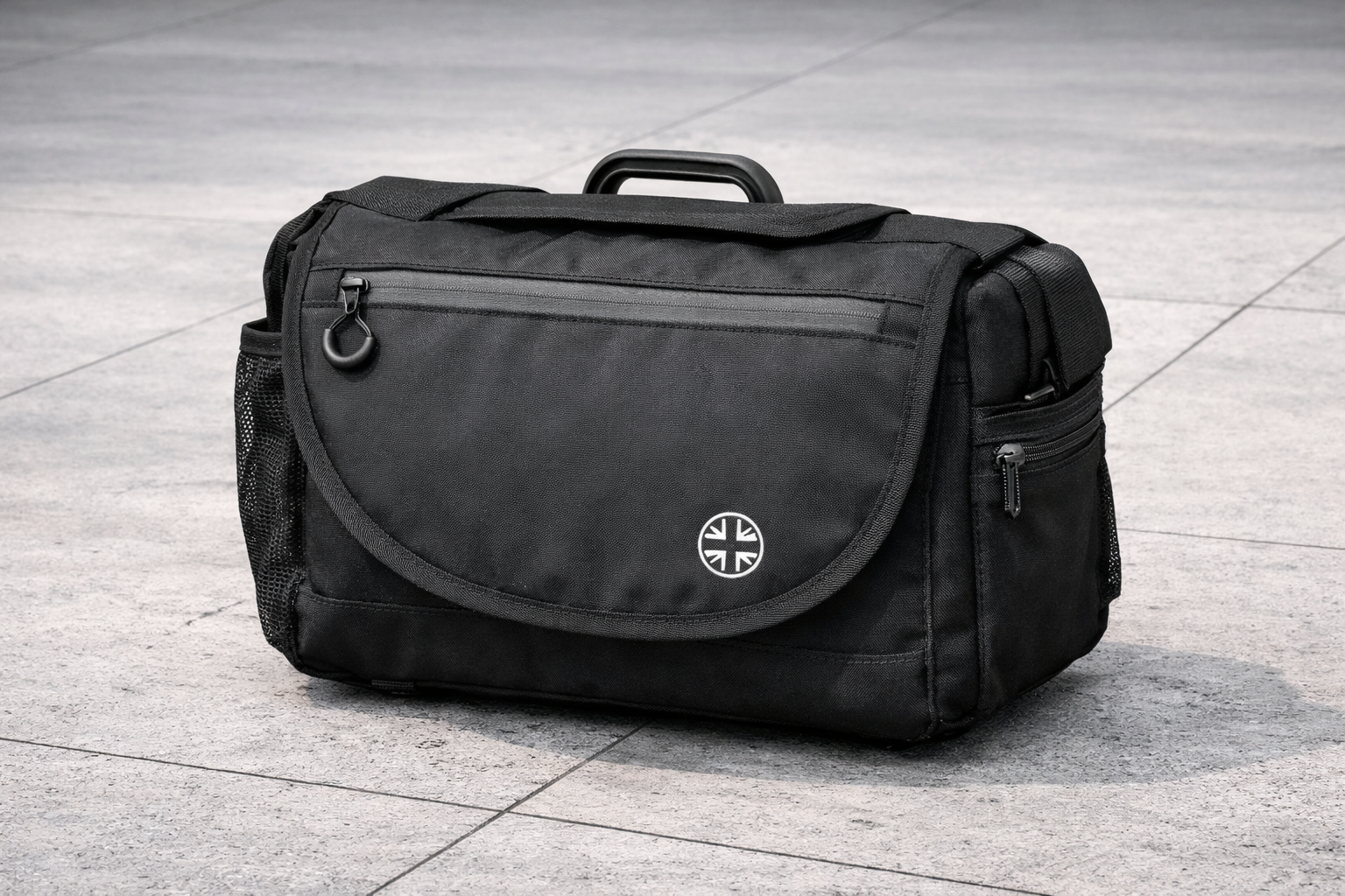 A black messenger bag with a front zipper pocket, side mesh pocket, and a round logo, placed on a light gray stone surface outdoors.