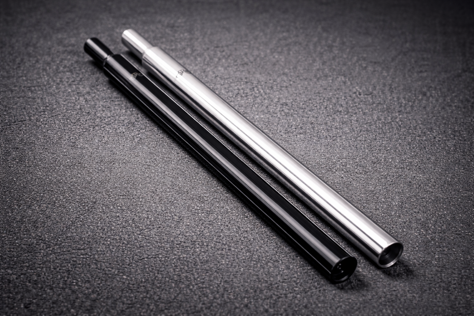 Two metal straws, one silver and one black, are placed side by side on a textured dark gray surface. The straws have a sleek, cylindrical design and appear shiny under the lighting.
