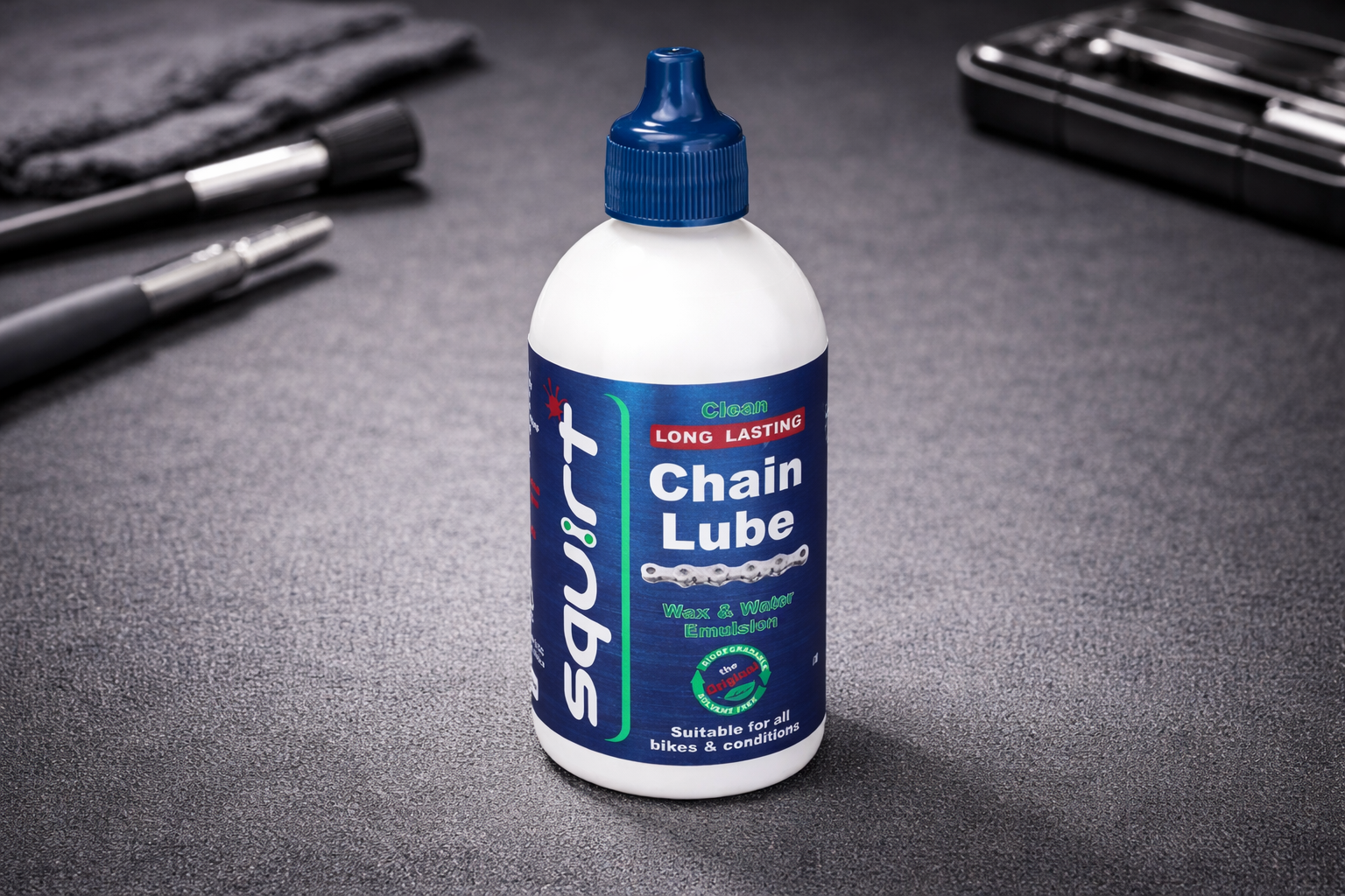 A white bottle of Squirt Chain Lube with a blue label stands upright on a gray surface, surrounded by bike maintenance tools in the background.