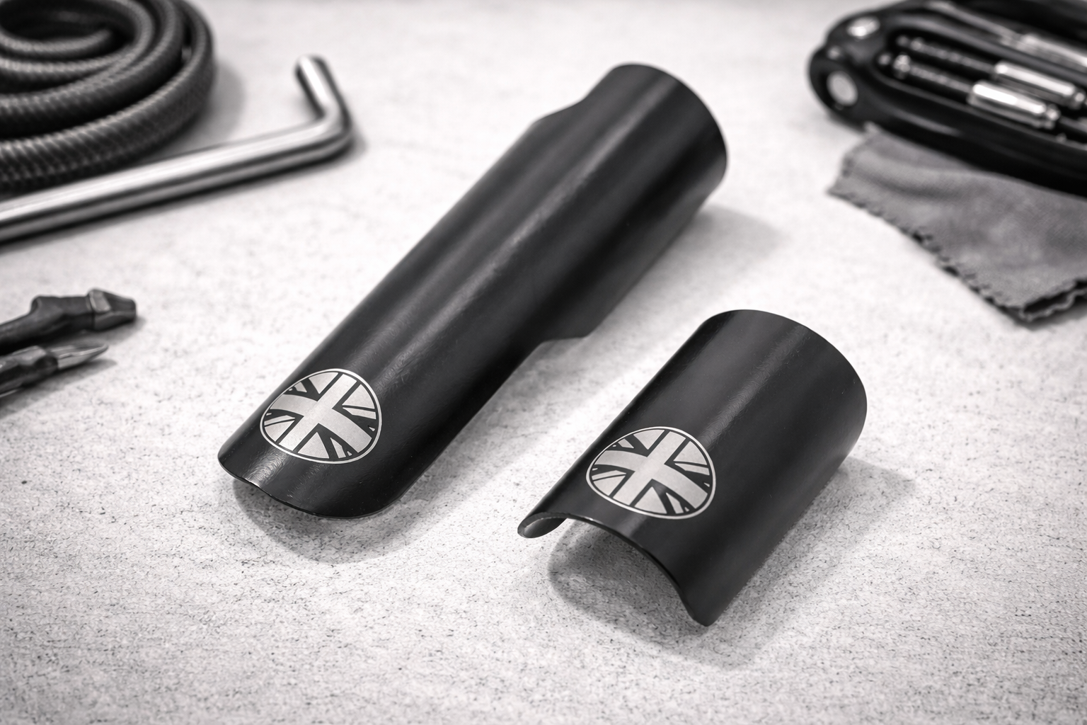 Two black metal covers with Union Jack emblems lie on a light surface, surrounded by tools including pliers, a hex key, a coiled cable, and a multitool with a cloth.