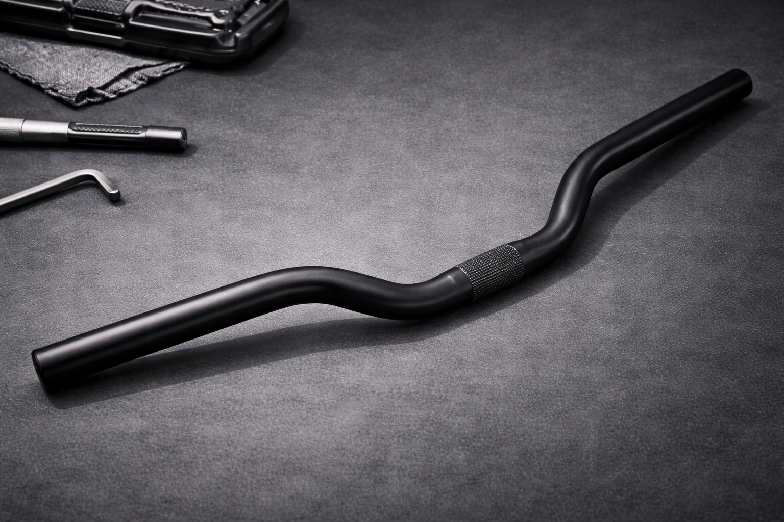 A black bicycle handlebar with a textured grip section is displayed on a dark surface, surrounded by various tools and metallic objects.