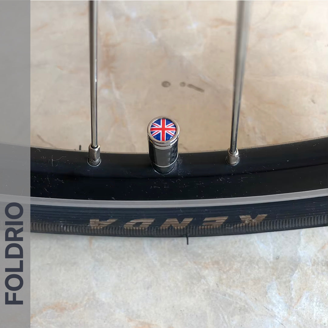Close-up of a bicycle wheel with a KENDA tire, fitted with FOLDRIO Brompton Union Jack Valve Caps – Schrader (A/C/G Line); "FOLDRIO" appears vertically on the left side of the image.