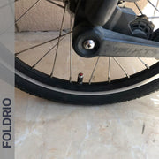 Close-up of a bicycle wheel with tire, spokes, and FOLDRIO Brompton Union Jack Valve Caps – Schrader (A/C/G Line). Tiled floor and beige wall in the background; "FOLDRIO" is written vertically on a gray stripe to the left.