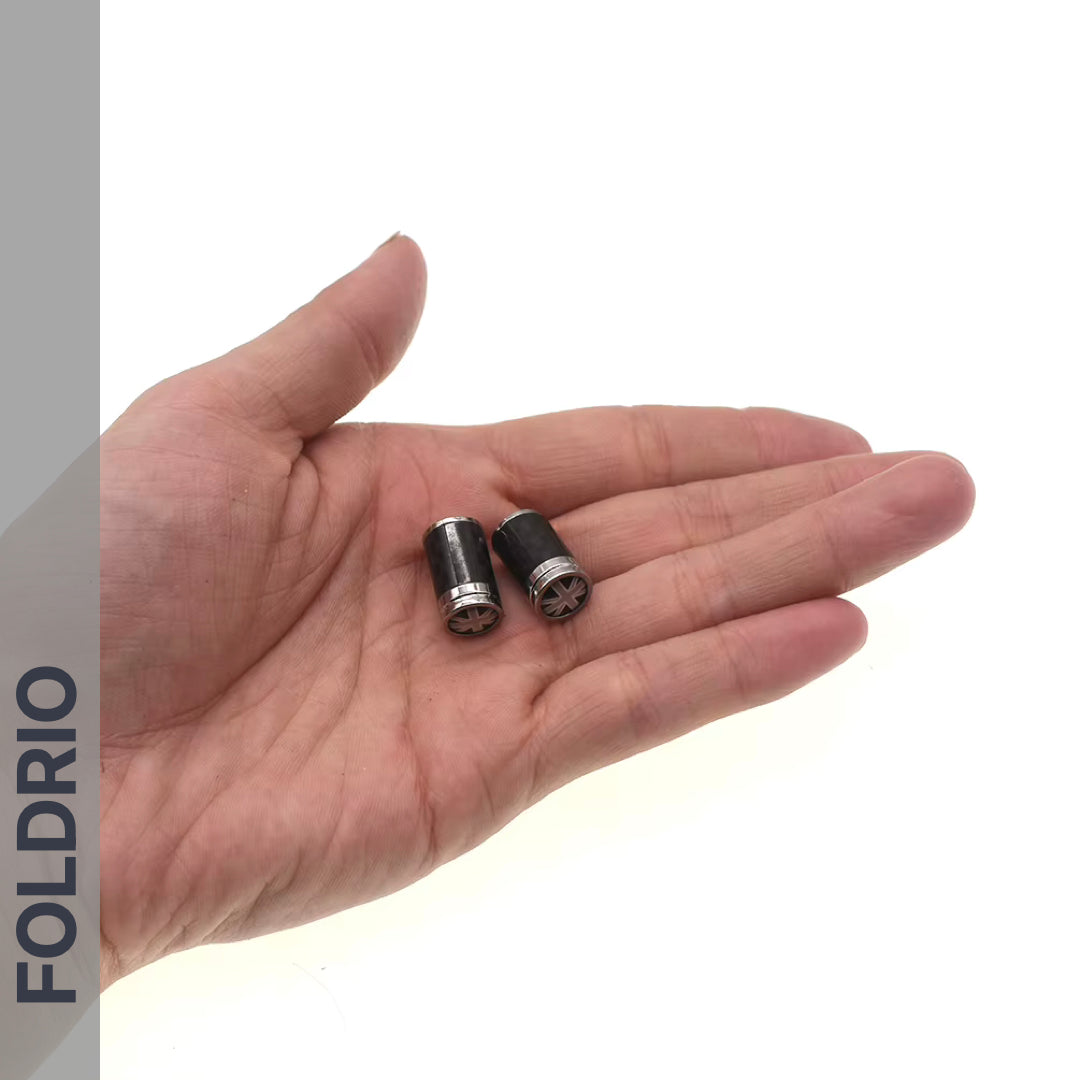 A hand holds two small, cylindrical Brompton Union Jack Valve Caps – Schrader (A/C/G Line) by FOLDRIO on the palm. The word "FOLDRIO" appears vertically on the left, with a plain white background.
