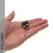 A hand holds two small cylindrical Brompton Union Jack Valve Caps – Schrader (A/C/G Line) by FOLDRIO, featuring red and black designs. "FOLDRIO" appears vertically on a gray strip along the left side.