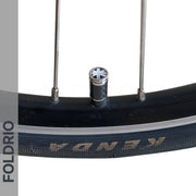 Close-up of a bicycle wheel with a black Kenda tire, two metal spokes, and a FOLDRIO Brompton Union Jack Valve Cap – Schrader (A/C/G Line). A gray vertical bar on the left reads FOLDRIO; carbon and copper accents add subtle detail.