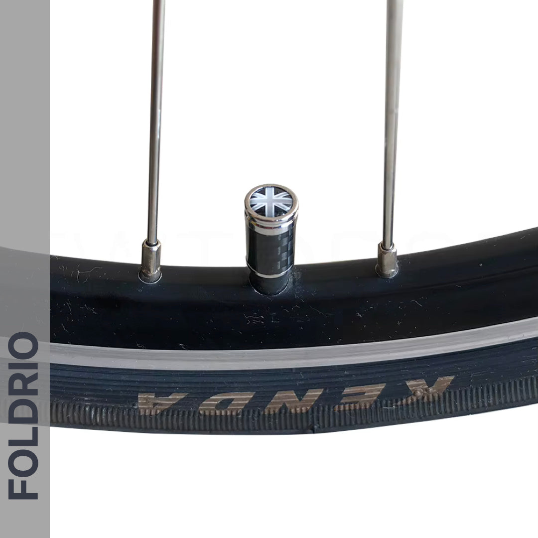 Close-up of a bicycle wheel with a black Kenda tire, two metal spokes, and a FOLDRIO Brompton Union Jack Valve Cap – Schrader (A/C/G Line). A gray vertical bar on the left reads FOLDRIO; carbon and copper accents add subtle detail.
