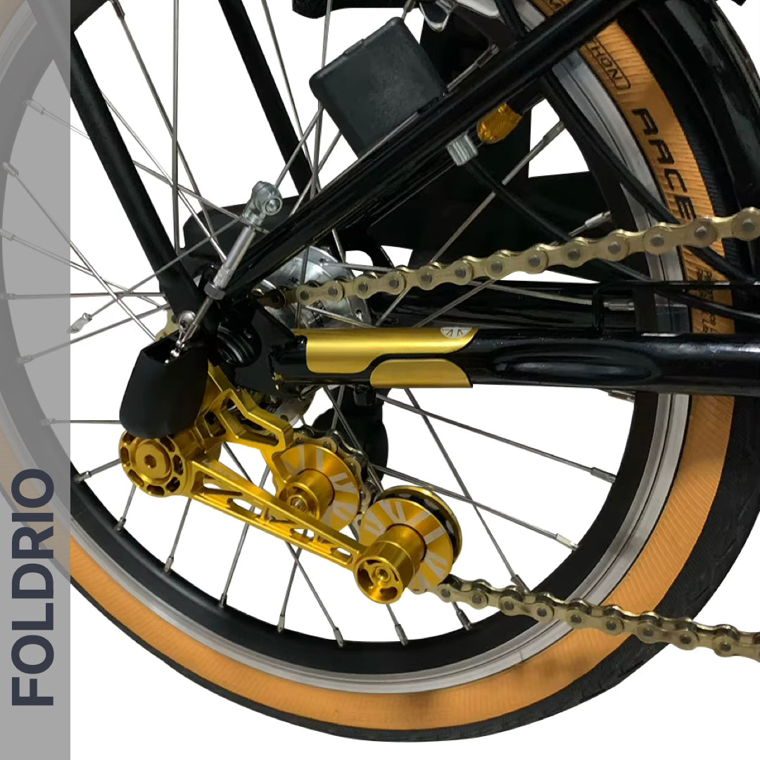 Close-up of a bicycle’s rear wheel with gold derailleur, tan sidewall tire, gold bike chain, and black frame featuring the FOLDRIO Brompton Frame Protector Set with British Flag. The FOLDRIO logo appears vertically on the left.