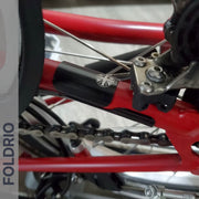 Close-up of a red bicycle's rear wheel, gear, and chain with a small black and white circular logo, plus a Brompton Frame Protector Set with British Flag by FOLDRIO. "FOLDRIO" appears vertically on the left edge.