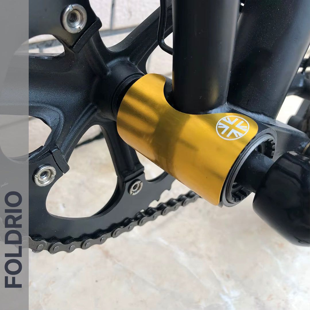 Close-up of a bicycle crankset with a gold bottom bracket and silver emblem, plus part of the chainring and pedal. A FOLDRIO Brompton Frame Protector Set with British Flag adds durability. The word FOLDRIO appears along the left edge.