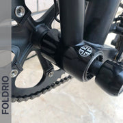 Close-up of a bicycle’s crankset and bottom bracket, showing a black frame with a white logo. A FOLDRIO Brompton Frame Protector Set with British Flag appears on the frame, with "FOLDRIO" printed vertically on a translucent band at the left.