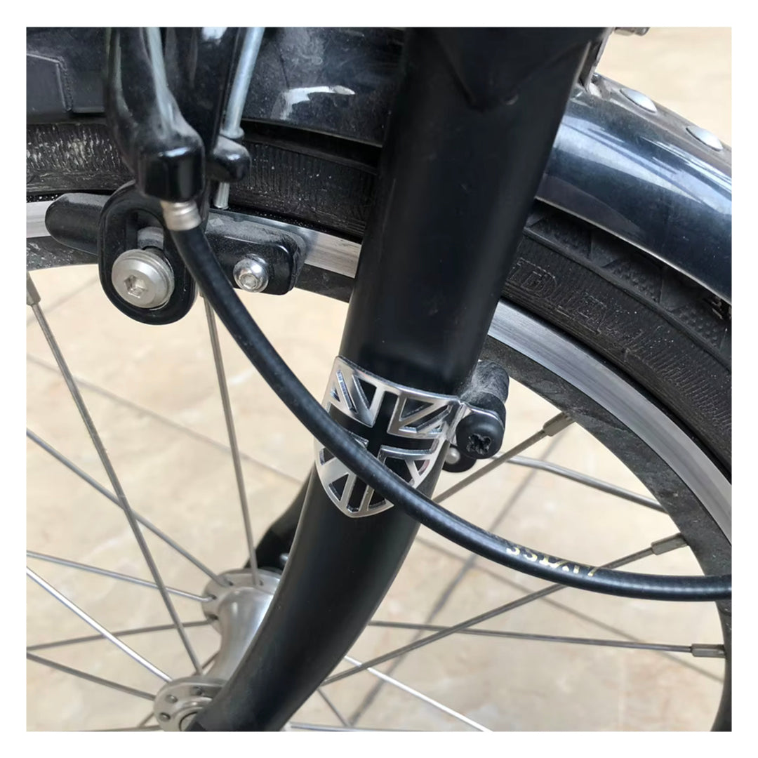 Close-up of a FOLDRIO Brompton Frame Protector Set with British Flag, fitted on a black bicycle front fork. Visible spokes, brake caliper, tire, and the protector are shown against a light-colored paved surface.