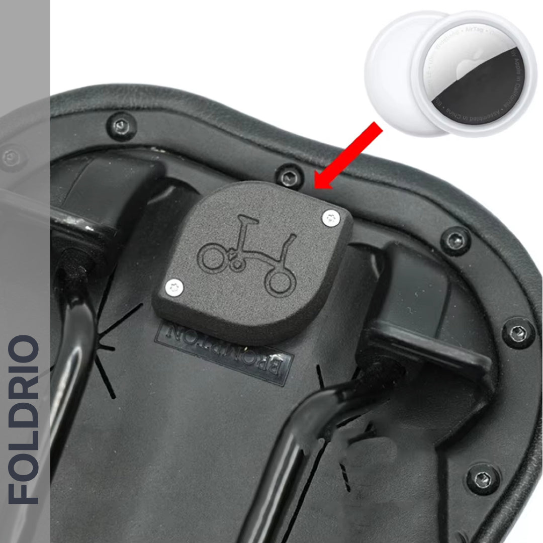 Close-up of the Brompton AirTag Saddle Mount, a black bike saddle underside with a hidden tracker holder. An inset shows a white AirTag with red arrow pointing to the holder. FOLDRIO branding highlights this anti-theft bike tracker.