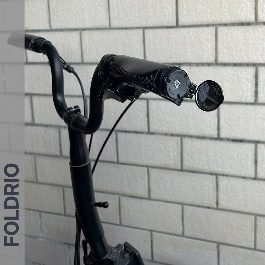 A close-up of a black electric scooter handlebar with a Brompton Bar End Mirror – 360° Adjustable, Foldable, set against a white brick wall. The word FOLDRIO is displayed vertically on the left side of the image.