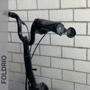 A close-up of a black electric scooter handlebar with a Brompton Bar End Mirror – 360° Adjustable, Foldable, set against a white brick wall. The word FOLDRIO is displayed vertically on the left side of the image.