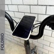 A Brompton Phone Mount – Lightweight, No Case Needed by FOLDRIO securely holds a smartphone on bicycle handlebars in front of a light-colored brick wall. The word FOLDRIO appears vertically on the left side of the image.