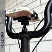 A smartphone is mounted on a bicycle’s handlebars using the FOLDRIO Brompton Phone Mount—lightweight and fits without a case. “FOLDRIO” appears vertically on the left against a white tiled wall in the background.