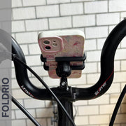 A Brompton Phone Mount by FOLDRIO, lightweight and requiring no case, secures a smartphone on a black bicycle’s handlebars. The word FOLDRIO appears vertically at the left in front of a white brick wall.