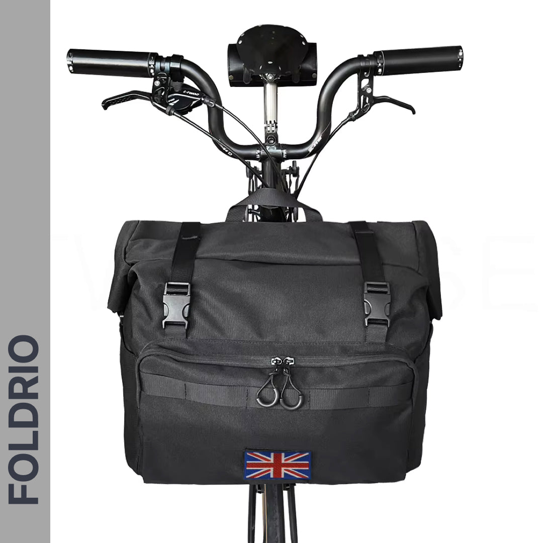 A black FOLDRIO Brompton Roll-Top Front Bag (27.5L) with buckle closures and a Union Jack patch is mounted on the handlebars via a front carrier block. The image features a gray banner with "FOLDRIO" text.