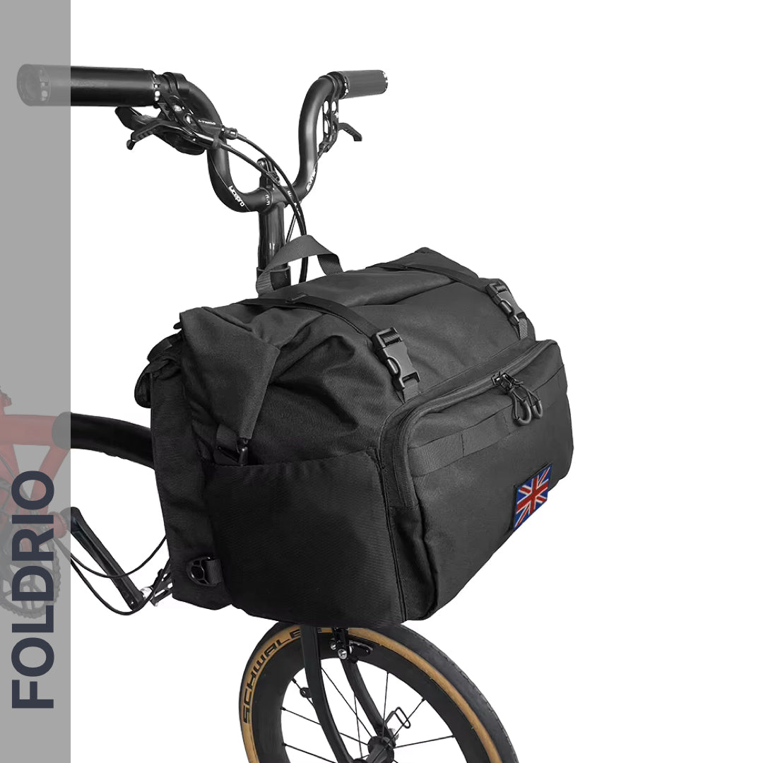 A black bicycle with a Brompton Roll-Top Front Bag – 27.5L by FOLDRIO is shown, the bag features a small Union Jack patch and is attached to the handlebars, set against a white background with "FOLDRIO" along the left side.