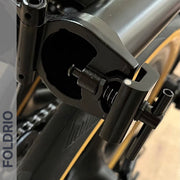 Close-up of a Brompton Stem Fixing Buckle Spring by FOLDRIO, showing the black frame hinge mechanism with a partially open locking clamp. The word FOLDRIO appears vertically on the left side of the image.