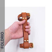 A hand holds the FOLDRIO Brompton Leather Carry Handle, made from brown full-grain leather with brass hardware, against a white background. The word FOLDRIO appears vertically on the left side.
