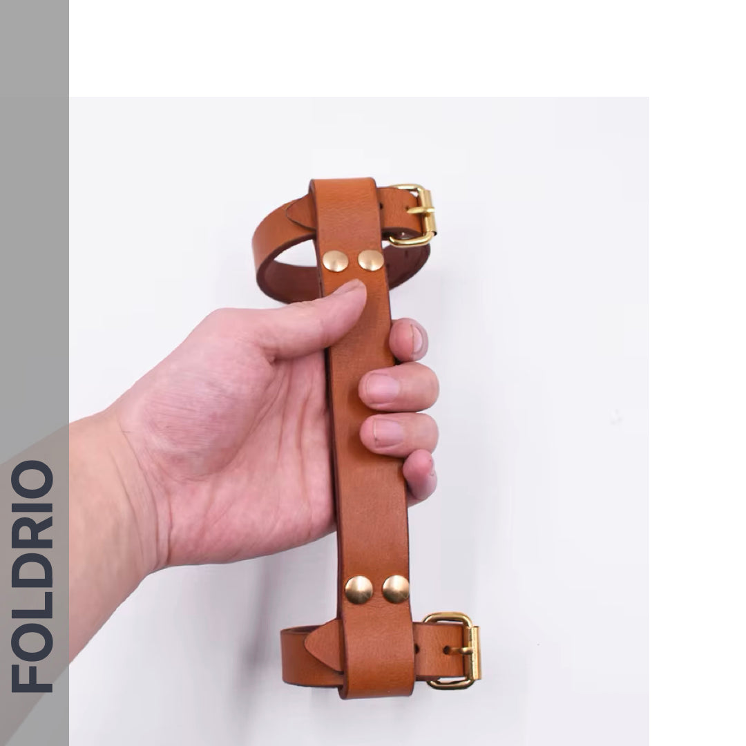 A hand holds the FOLDRIO Brompton Leather Carry Handle, made from brown full-grain leather with brass hardware, against a white background. The word FOLDRIO appears vertically on the left side.