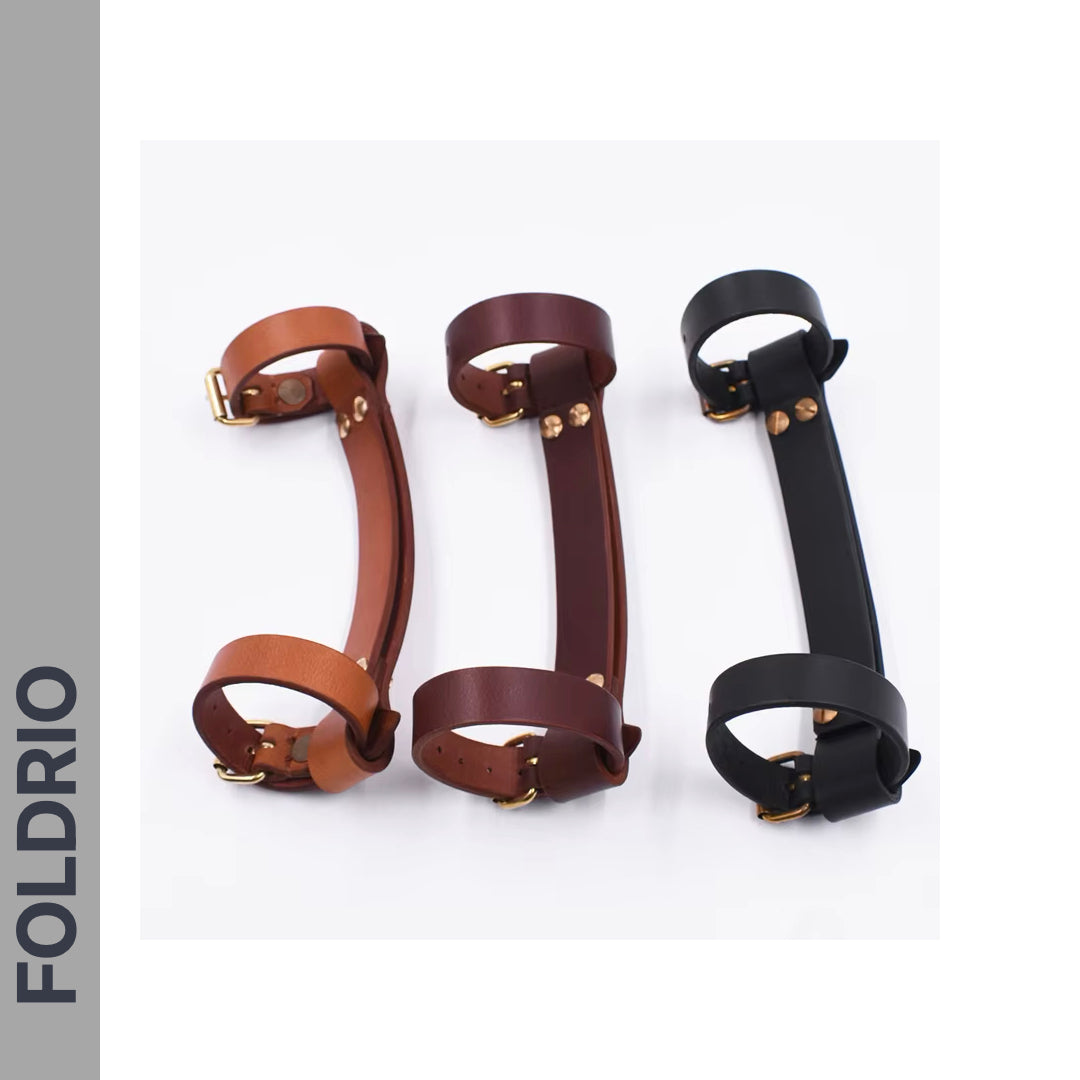 Three Brompton Leather Carry Handles by FOLDRIO, crafted from full-grain leather with brass hardware in brown, dark brown, and black, are displayed side by side. "FOLDRIO" appears vertically on a gray strip to the left.
