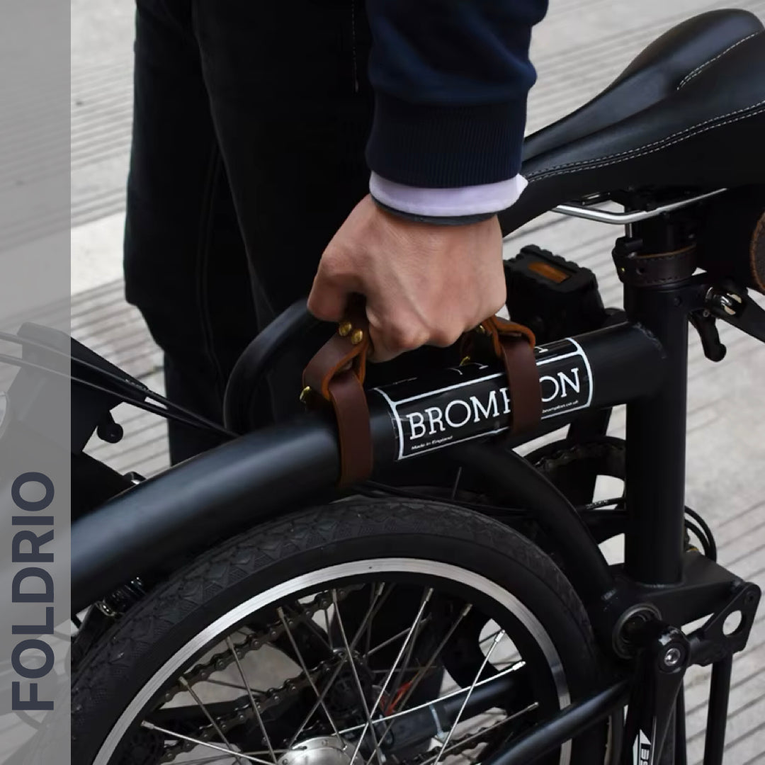 A person grips the FOLDRIO Brompton Leather Carry Handle – Full-Grain, Brass Hardware, attached to a folded Brompton bike frame. The word FOLDRIO appears vertically on the image’s left side.
