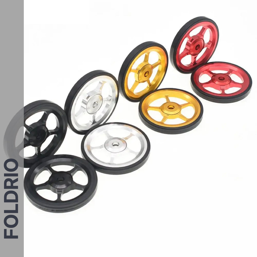 Eight FOLDRIO Brompton Easy Wheels with Titanium Bolts—three-spoke, lightweight aluminium in black, silver, gold, and red—are displayed in two curved rows on white. The word FOLDRIO appears vertically on the left side.