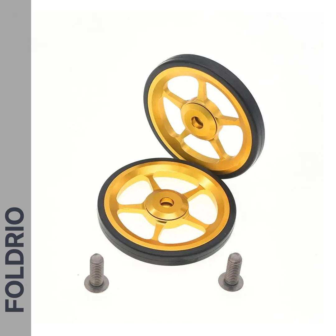 Two gold and black metal FOLDRIO Brompton Easy Wheels with titanium bolts, each featuring four spokes, are shown on a white background with two silver screws below. The FOLDRIO brand name appears vertically on the left side.