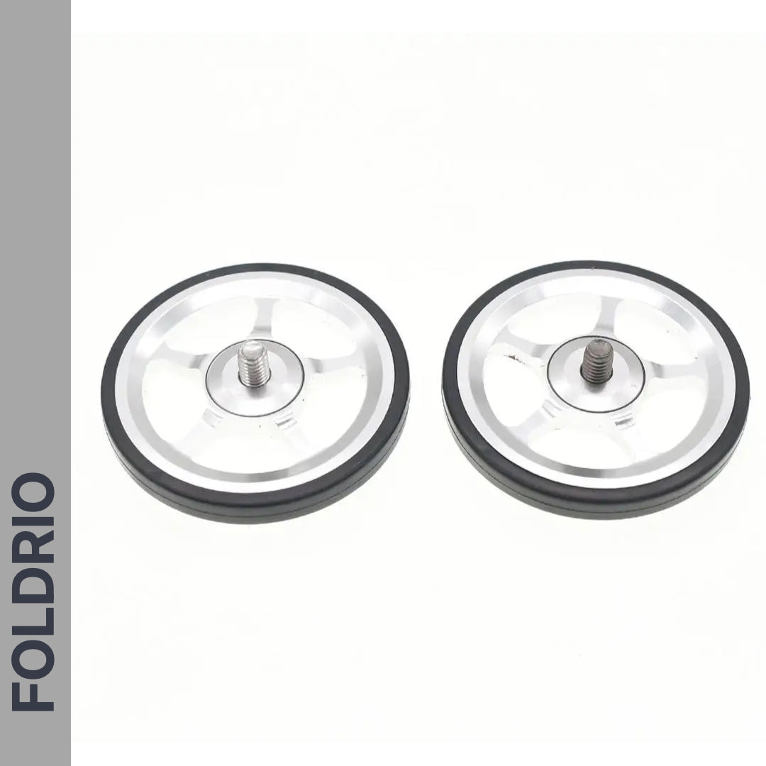 Two silver and black Brompton Easy Wheels with titanium bolts by FOLDRIO are placed side by side on a white background, with the FOLDRIO logo displayed vertically on a gray strip along the left edge.