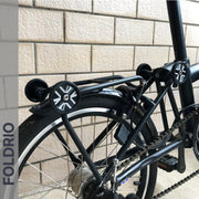 Close-up of a FOLDRIO folding bike’s rear section with Brompton Easy Wheels Union Jack Edition (titanium bolts), rear rack, chain, and small tire—all set against a brick wall backdrop.