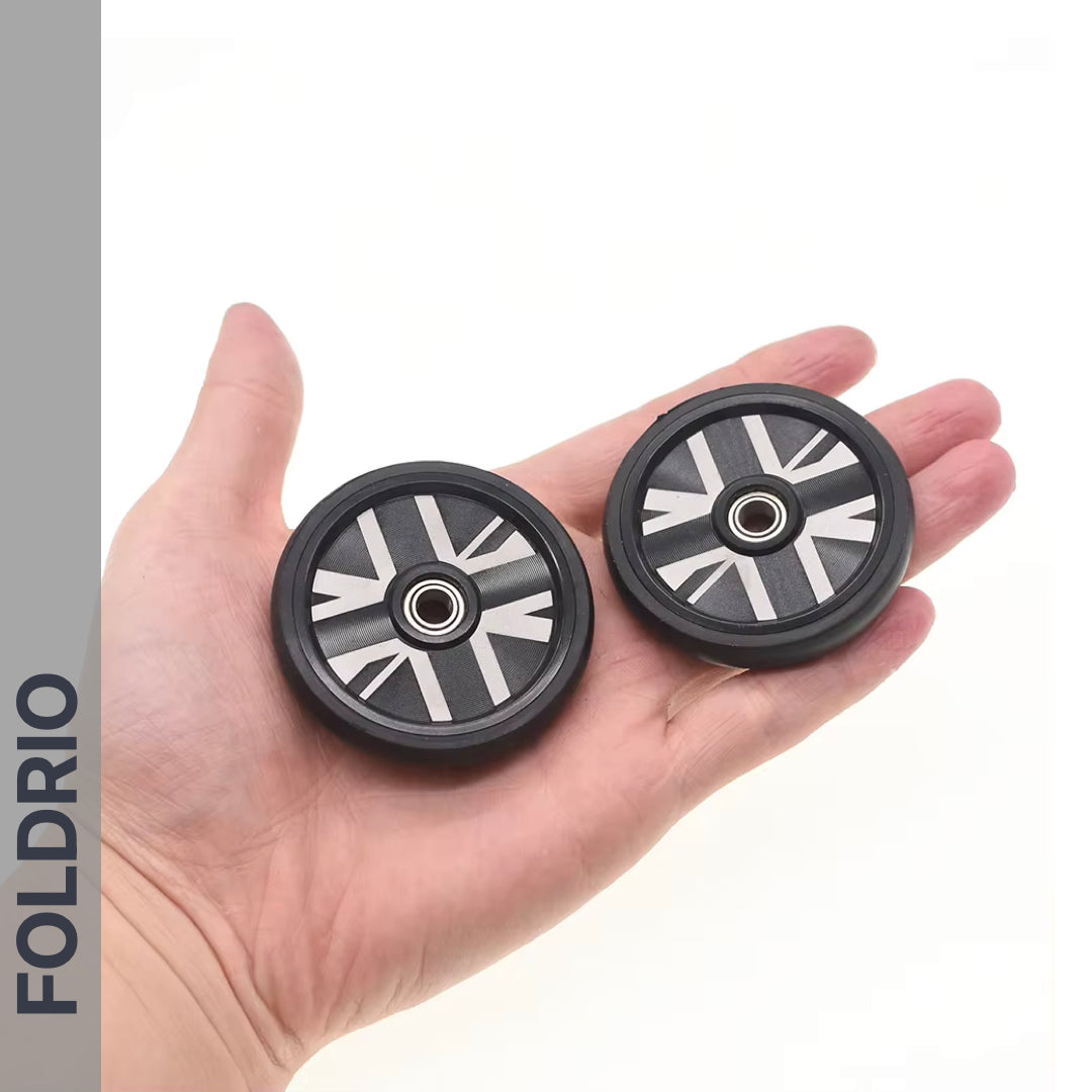 A hand holds two FOLDRIO Brompton Easy Wheels Union Jack Edition with titanium bolts—sleek black wheels featuring silver geometric patterns, central bearings, and the brand name vertically displayed on the left.