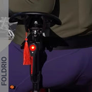 Close-up of a person in dark clothing sitting on a compact, foldable electric scooter equipped with the Brompton Smart Brake Tail Light by FOLDRIO. The brand name FOLDRIO appears vertically on the scooter's left side.