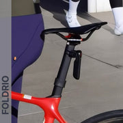 Close-up of a red road bike saddle and seat post with the FOLDRIO Brompton Smart Brake Tail Light, featuring an intelligent brake sensor. A person in purple leggings stands nearby; another in white shoes is in the background.
