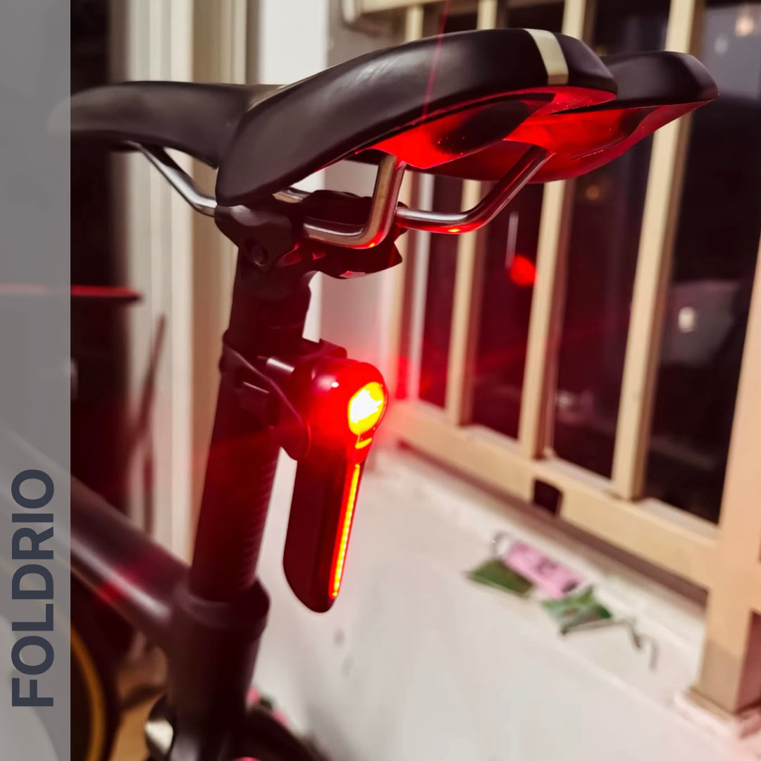 A close-up of a Brompton Smart Brake Tail Light by FOLDRIO glowing bright red on a bike saddle mount, with the bicycle indoors near a window and FOLDRIO vertically displayed on the left side of the image.