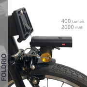 Close-up of the FOLDRIO Brompton Front Fork Light (400 lumens) in black, mounted above the front tire. Features an adjustable beam, 2000 mAh capacity, USB-C rechargeability, and FOLDRIO branding vertically on the left side.