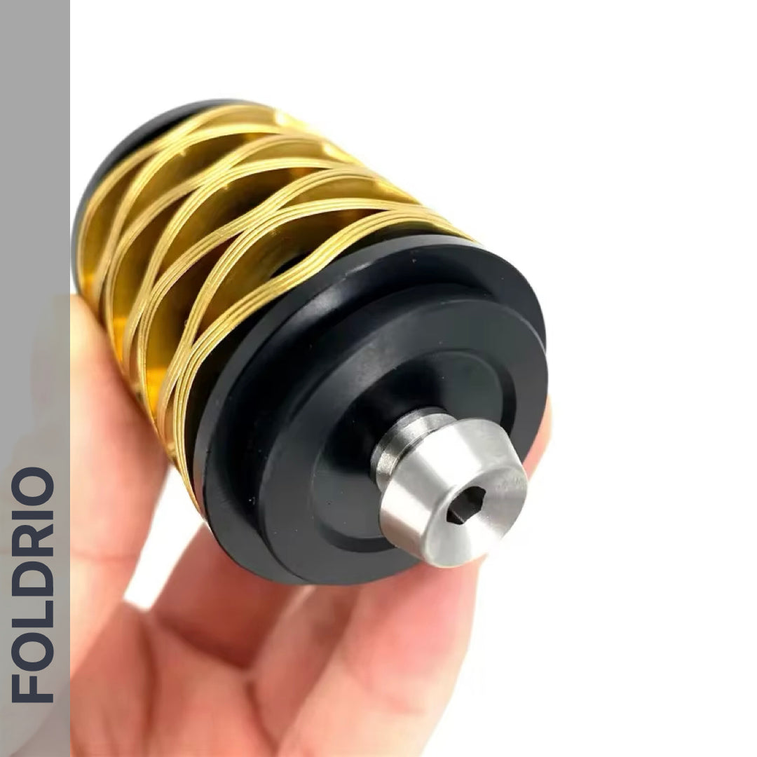 A hand holds the Brompton Rear Shock Absorber by FOLDRIO—a cylindrical object with a black base, metallic silver tip, and gold wire coil. The brand name FOLDRIO is displayed vertically on the left side.