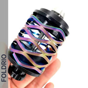 A hand holds a Brompton Rear Shock Absorber by FOLDRIO, a black cylindrical part wrapped with iridescent coiled wires; "FOLDRIO" appears vertically on the left side of the image.