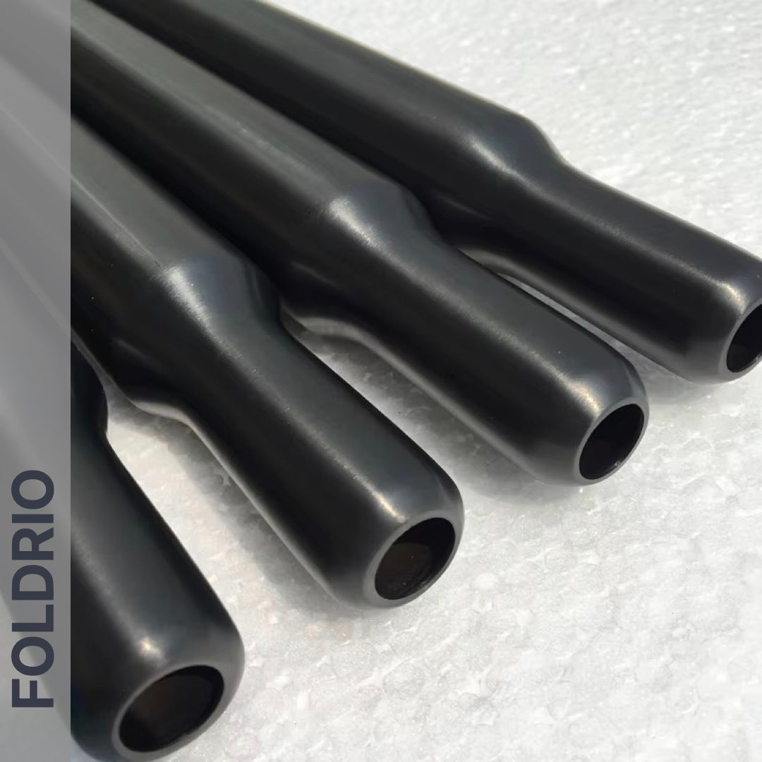 Four multicolour titanium Brompton seatposts from FOLDRIO are laid parallel on a textured white surface, with the brand name FOLDRIO shown vertically on the left.