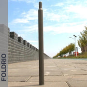 A Brompton Titanium Seatpost – Multicolour Edition by FOLDRIO stands upright on a paved sidewalk beside a stone wall and trees under a blue, partly cloudy sky. The brand name FOLDRIO appears vertically on the left side.