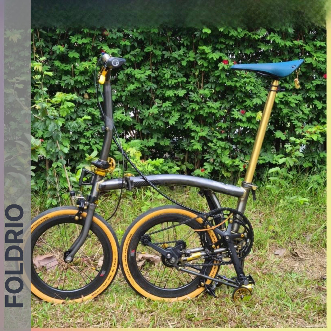 A Brompton Titanium Seatpost – Multicolour Edition by FOLDRIO stands on grass before a leafy hedge, with "FOLDRIO" written vertically on the image’s left side. The bicycle features gold and black detailing and a compact folding design.