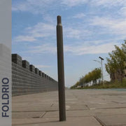 A Brompton Titanium Seatpost – Multicolour Edition by FOLDRIO stands upright on a paved sidewalk next to a stone wall and trees under a partly cloudy sky. The brand name FOLDRIO is vertically written on the left side.