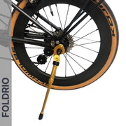Close-up of a bicycle’s rear wheel with the FOLDRIO Brompton Kickstand – Adjustable Aluminium (A and C Line) extended; the tire features a tan sidewall, bold black lettering, and "FOLDRIO" displayed vertically on the left.
