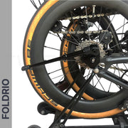 Close-up of a folded bicycle with gold-rimmed tires and black spokes, featuring the FOLDRIO Brompton Kickstand – Adjustable Aluminium (A and C Line) mounted near the rear axle. "FOLDRIO" is displayed vertically on a gray strip to the left.