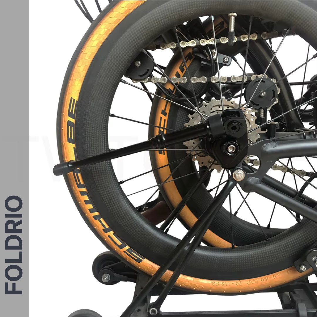 Close-up of a folded bicycle with gold-rimmed tires and black spokes, featuring the FOLDRIO Brompton Kickstand – Adjustable Aluminium (A and C Line) mounted near the rear axle. "FOLDRIO" is displayed vertically on a gray strip to the left.