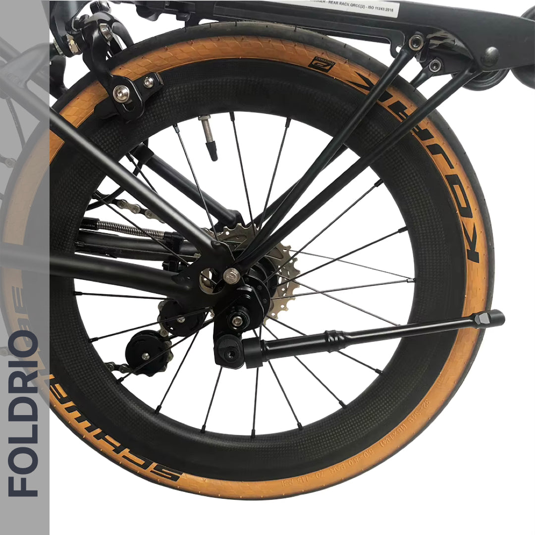 Close-up of a folded bicycle wheel with tan sidewalls, spokes, and gear mechanism. The FOLDRIO Brompton Kickstand – Adjustable Aluminium, A and C Line rear axle mount is visible. "FOLDRIO" is shown vertically on a gray bar at the left.