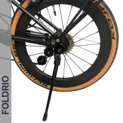 Close-up of a bike's rear wheel with tan sidewall tire, visible gears, rear axle mount, and an extended Brompton Kickstand – Adjustable Aluminium (A & C Line) by FOLDRIO. The word FOLDRIO appears vertically on the left.