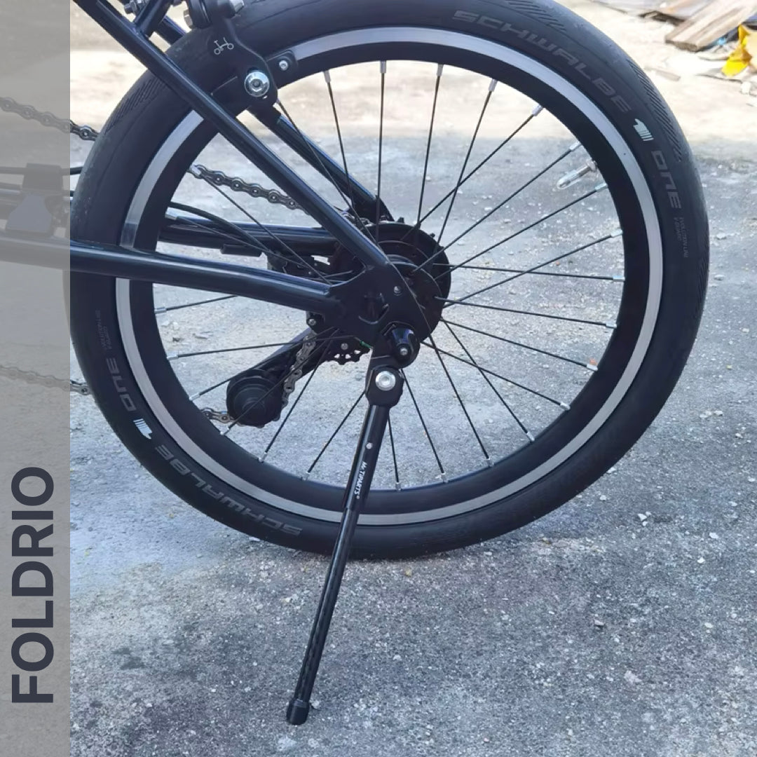 Close-up of a Brompton Kickstand – Carbon Fibre, P and T Line by FOLDRIO, extended on concrete under a bike’s rear wheel with a Schwalbe One tire; “FOLDRIO” appears vertically on the left side of the image.
